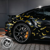 Black car with gold bat decals parked on a brick road, with a logo in the corner.