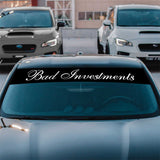 Car with 'Bad Investments' decal on the windshield in a parking lot.
