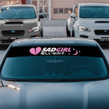Car with a 'SAD GIRL' decal on the windshield in a parking lot.