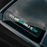Car window with a decal reading 'TITS ARE Real SMILE IS Fake' and a logo at the bottom.
